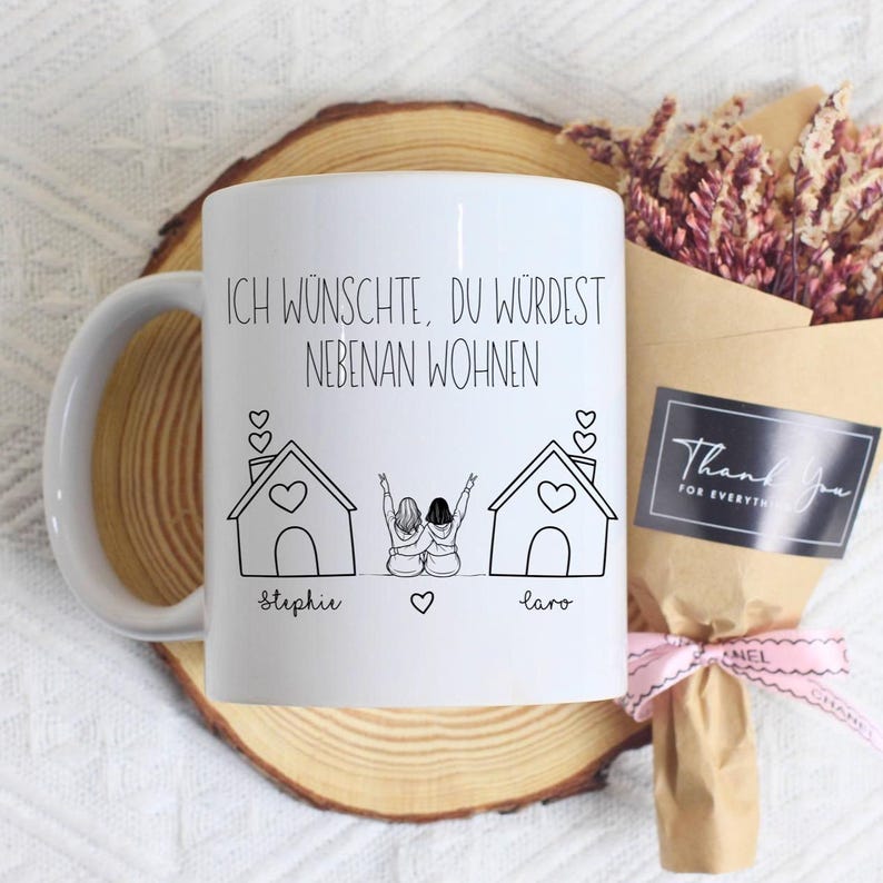 Puede incluir: Taza de cer&aacute;mica blanca con una ilustraci&oacute;n negra de dos casas y dos figuras con los brazos levantados. La taza tiene el texto "ICH W&Uuml;NSCHTE, DU W&Uuml;RDEST NEBENAN WOHNEN" y los nombres "Stephie" y "Laro". Un ramo de flores secas y una tarjeta "Thank You" est&aacute;n al lado.