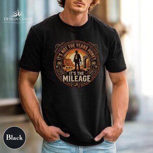 K&ouml;nnte beinhalten: Schwarzes T-Shirt mit einem kreisf&ouml;rmigen Grafikdesign, das eine Silhouette eines Mannes mit Hut und Peitsche zeigt, mit dem Text "IT'S NOT THE YEARS HONEY IT'S THE MILEAGE". Das Design umfasst W&uuml;stenlandschaft und einen Seilrand.