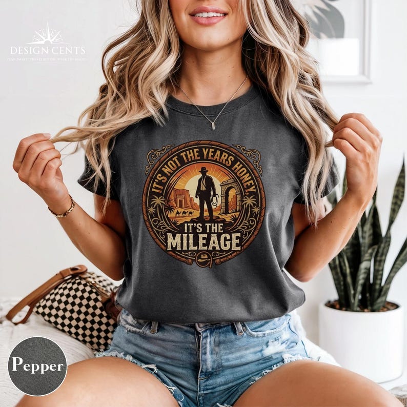 K&ouml;nnte beinhalten: Dunkelgraues T-Shirt mit einem kreisf&ouml;rmigen Grafikdesign. Die Grafik zeigt eine Silhouette einer Person mit Hut, die eine Peitsche h&auml;lt, mit dem Text "IT'S NOT THE YEARS HONEY IT'S THE MILEAGE". Das Design ist in Braun- und Oranget&ouml;nen gehalten.