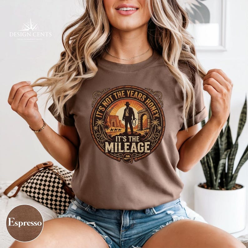 K&ouml;nnte beinhalten: Espressofarbenes T-Shirt mit einem kreisf&ouml;rmigen Grafikdesign, das einen Cowboy, eine W&uuml;stenlandschaft und den Text "IT'S NOT THE YEARS HONEY IT'S THE MILEAGE" zeigt. Das Design ist in Braun-, Beige- und Oranget&ouml;nen gehalten. Das T-Shirt hat kurze &Auml;rmel.