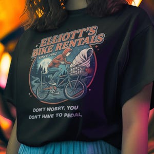 May include: Black t-shirt featuring a graphic of a person riding a bicycle with a large ice cream cone in the basket. The text "ELLIOTT'S BIKE RENTALS SINCE 1982" and "DON'T WORRY, YOU DON'T HAVE TO PEDAL" are printed on the shirt.