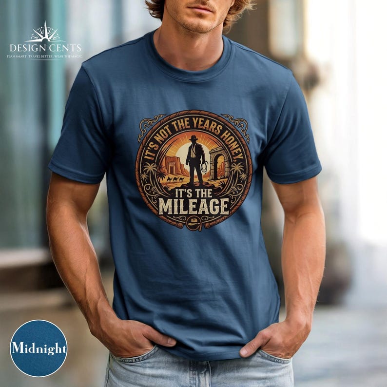 K&ouml;nnte beinhalten: Nachtblaues T-Shirt mit einem kreisf&ouml;rmigen Grafikdesign, das eine Silhouette eines Mannes mit Hut und Peitsche zeigt, mit dem Text "IT'S NOT THE YEARS HONEY IT'S THE MILEAGE". Das Shirt hat auch das Design Cents Logo.