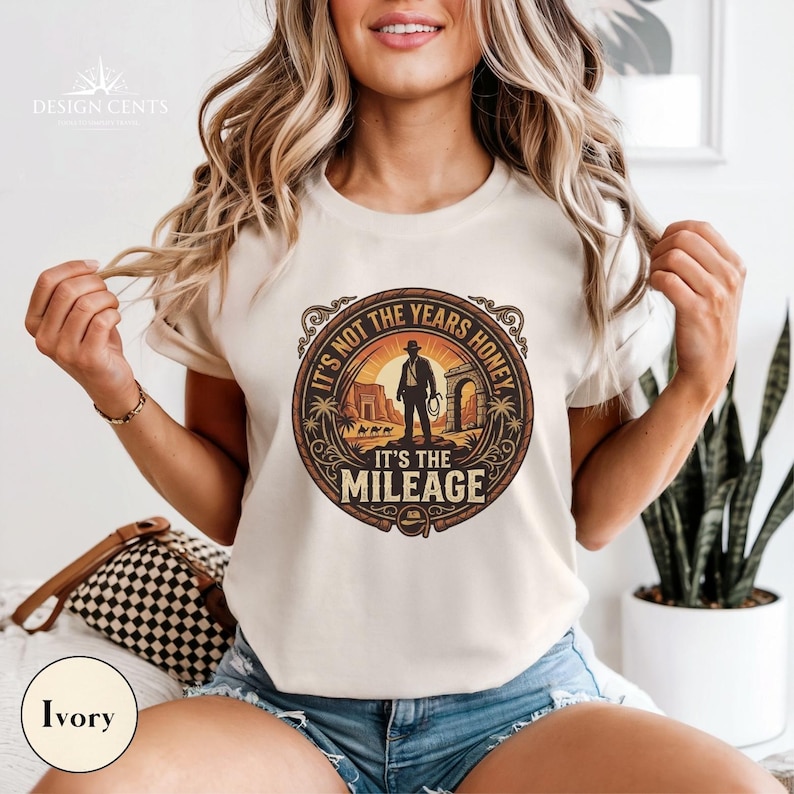 K&ouml;nnte beinhalten: Elfenbeinfarbenes T-Shirt mit einem kreisf&ouml;rmigen Aufdruck. Der Aufdruck zeigt eine Silhouette einer Person mit Hut, eine W&uuml;stenlandschaft und den Text "IT'S NOT THE YEARS HONEY, IT'S THE MILEAGE". Das T-Shirt hat kurze &Auml;rmel.
