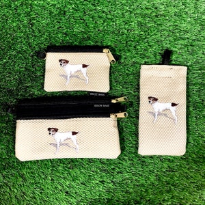 May include: Three beige zippered pouches with black trim and a white Jack Russell Terrier dog embroidered on each. The pouches are on a green grass background. The pouches have the text "Dixie Bags" on the bottom.