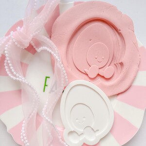 Peut inclure: Une assiette à rayures roses et blanches avec un moule rose en forme d'œuf et un moule blanc en forme d'œuf. Un ruban rose avec des perles est drapé sur l'assiette. Le moule rose a un motif d'œuf souriant.