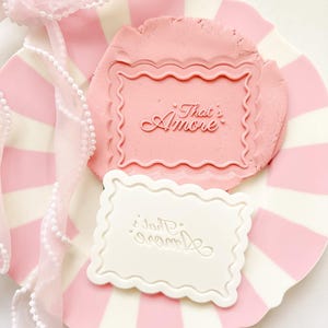 May include: A pink and white striped plate with a pink clay stamp that says "That's Amore" and a white stamp with the same text. A sheer pink ribbon with pearls is draped over the plate.