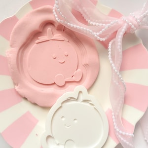 Peut inclure: Une assiette à rayures roses et blanches contient une empreinte en argile rose d'un personnage de dessin animé souriant. Un moule en plastique blanc du même personnage est à proximité. Un ruban rose transparent avec des perles est noué en nœud.