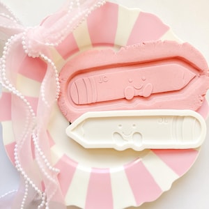 Peut inclure: Une assiette à rayures roses et blanches présente un moule en forme de crayon rose et un emporte-pièce en forme de crayon blanc, tous deux avec des visages souriants et les initiales "JC". Un ruban rose avec des perles blanches est drapé sur l'assiette.