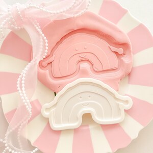 Peut inclure: Une assiette à rayures roses et blanches présente un moule en forme d'arc-en-ciel rose et un emporte-pièce en forme d'arc-en-ciel blanc. Un ruban rose à bordure perlée est noué en un nœud, créant une présentation fantaisiste sur le thème pastel.