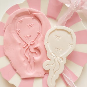Puede incluir: Un molde rosa con forma de globo y un objeto blanco con forma de globo con detalles de lazo se encuentran sobre un plato a rayas rosas y blancas. Una cinta rosa con perlas está drapeada sobre el plato. El molde está prensado en arcilla rosa.