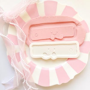 Peut inclure: Une assiette à rayures roses et blanches contient un rectangle d'argile rose avec un motif de règle et un moule en forme de règle blanc. Un ruban rose transparent avec une garniture de perles blanches est drapé sur l'assiette. Les motifs de règles ont des visages souriants.