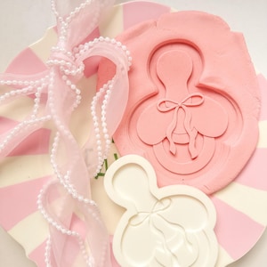May include: A pink and white striped plate with a pink ribbon and a white beaded trim. A pink clay mold of a baby pacifier with a bow is next to a white pacifier mold. The image has a soft, feminine aesthetic.