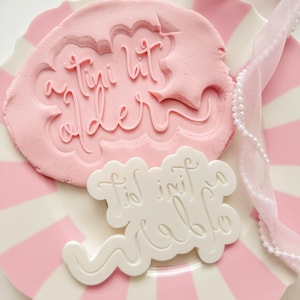 May include: A pink clay with the words "a tiny bit older" pressed into it, and a white plastic mold with the words "tid ini table" on a pink and white striped plate. A white ribbon with pearls is on the plate.