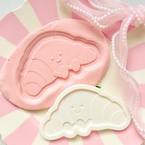 Peut inclure: Une assiette à rayures roses et blanches présente un moule rose en forme de croissant avec un visage souriant et un emporte-pièce blanc en forme de croissant. Un ruban rose avec des perles est drapé sur l'assiette.