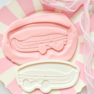 Peut inclure: Un emporte-pièce rose et un biscuit rose sur une assiette à rayures roses et blanches. L'emporte-pièce est blanc et en forme de camping-car. Un ruban rose avec des perles est drapé sur l'assiette.