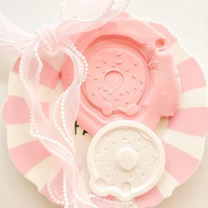 Peut inclure: Une assiette à rayures roses et blanches contient un moule en forme de donut rose et un objet en forme de donut blanc. Un ruban rose transparent avec une bordure perlée blanche est noué autour de l'assiette.