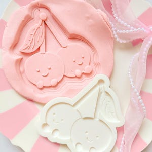 Peut inclure: Un emporte-pièce rose en forme de cerise et une empreinte en argile rose du même motif reposent sur une assiette à rayures roses et blanches. Un emporte-pièce blanc en forme de cerise est également présent. Un ruban rose avec des perles ajoute une touche décorative.