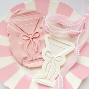 Op de afbeelding: Een roze en wit gestreepte plaat bevat een roze klei-afdruk en een witte plastic mal, beide in de vorm van een martiniglas met een strik. Een roze lint met parels is over de plaat gedrapeerd.