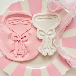 May include: A pink and white striped plate holds a pink clay impression and a white plastic mold of a champagne glass with a bow. A sheer, white ribbon with pearls is draped across the plate.