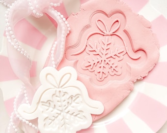 May include: A white and pink striped plate holds a pink dough with a snowflake bell design pressed into it. A white bell-shaped mold with a snowflake design and a pink ribbon with pearl accents are also present. The theme is holiday baking.