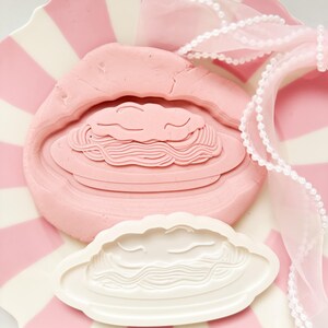 Peut inclure: Un moule rose en forme de nuage avec un motif de pâtes, à côté d'un moule blanc avec le même motif. Les moules reposent sur une assiette à rayures roses et blanches, avec un ruban rose et une garniture de perles.
