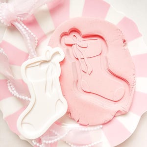 May include: A white stocking-shaped cookie cutter and a pink clay impression of a stocking with a bow, presented on a pink and white striped plate. Pink ribbon and a beaded trim enhance the festive holiday theme.
