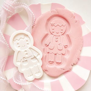 May include: A white gingerbread man cookie cutter and a pink gingerbread man impression on pink dough, displayed on a pink and white striped plate. A pink ribbon with white trim is draped around the plate, creating a festive, holiday theme.