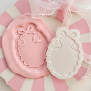 Puede incluir: Un plato a rayas rosas y blancas sostiene un molde rosa con un diseño de lazo y una etiqueta blanca con un diseño de lazo. Una cinta rosa transparente y cuentas de perlas están drapeadas sobre el plato, creando una estética delicada y femenina.