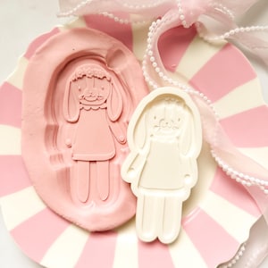 May include: A pink bunny-shaped mold and a white bunny-shaped object are displayed on a pink and white striped plate. A pink ribbon with a pearl strand is draped across the plate. The bunny has a floral crown and a dress.