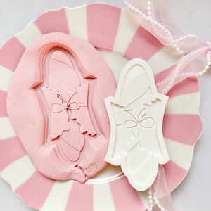 Peut inclure: Une assiette à rayures roses et blanches présente un moule rose et un emporte-pièce blanc, tous deux en forme de robe. Un ruban rose transparent avec une bordure de perles est drapé sur l'assiette.