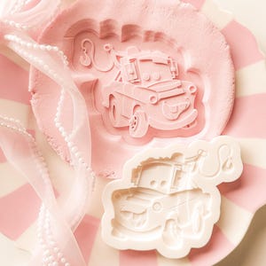 May include: A white plastic cookie cutter shaped like a tow truck with a hook and a smiling face, positioned next to a light pink clay impression of the same truck design, featuring detailed wheels, a front bumper, and a crane arm with a hook.
