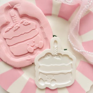 Peut inclure: Une assiette à rayures roses et blanches contient un moule à gâteau rose et un moule à gâteau blanc. Les moules à gâteau ont la forme d'un cupcake de dessin animé avec une bougie. Un ruban rose avec des perles blanches est en arrière-plan.