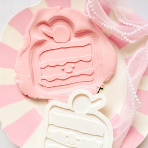 Peut inclure: Un moule rose en forme de gâteau et une empreinte en argile rose reposent sur une assiette à rayures roses et blanches. Un moule blanc en forme de gâteau est à côté de l'argile. Un ruban rose avec des perles blanches est drapé sur l'assiette.