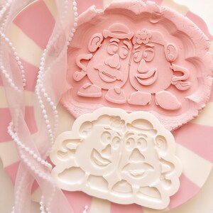 Könnte beinhalten: Ein weißer Kunststoff-Ausstecher mit den geprägten Gesichtern von Mr. und Mrs. Potato Head. Daneben liegt ein hellrosa Teigabdruck derselben Figuren. Ein transparentes rosa Band mit weißen Perlen liegt auf dem gestreiften rosa-weißen Teller.