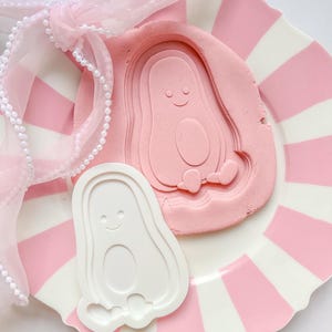 Peut inclure: Une assiette à rayures roses et blanches contient un moule rose en forme d'avocat et un emporte-pièce blanc en forme d'avocat. Un ruban rose avec des perles est noué autour de l'assiette. Les motifs d'avocats ont des visages souriants.