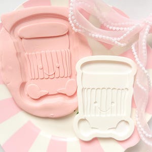 Peut inclure: Un emporte-pièce en forme de cupcake rose et un biscuit en forme de cupcake rose sur une assiette à rayures roses et blanches. Un ruban rose avec des perles blanches est sur l'assiette. Le découpoir et le biscuit ont un visage souriant.