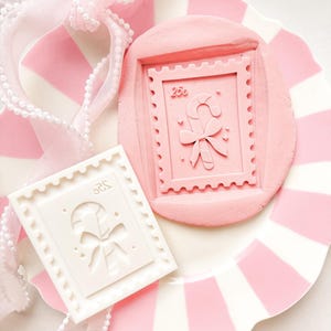 Puede incluir: Un cortador de galletas rosa con forma de sello y una galleta rosa con forma de sello sobre un plato a rayas rosas y blancas. El diseño del sello presenta un bastón de caramelo y un lazo. También hay un cortador de galletas blanco con forma de sello.
