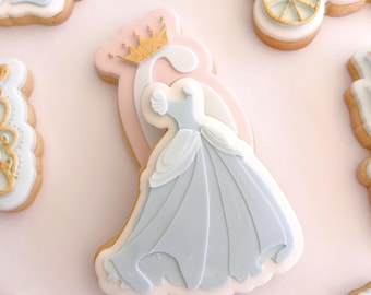 May include: A sugar cookie shaped like a ballgown in light blue and white icing, featuring a gold crown detail at the top. The cookie is decorated with layered icing to create the appearance of a dress, set against a light pink background.