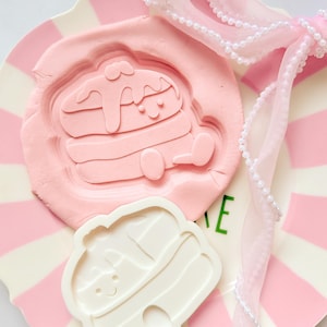 May include: A pink pancake-shaped mold and a white pancake-shaped cookie cutter sit on a pink and white striped plate. A pink ribbon with pearl accents is tied in a bow. The pancake mold has a smiling face.