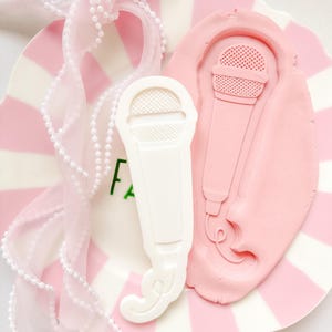 May include: A white microphone-shaped mold and a pink clay impression of a microphone rest on a pink and white striped plate. A pink ribbon with pearl accents is draped nearby. The word "FA" is visible on the plate.
