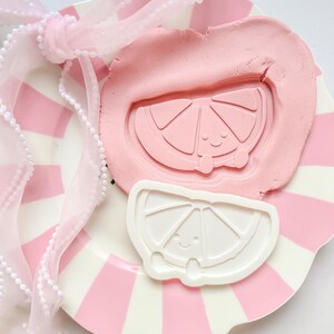 Peut inclure: Une assiette à rayures roses et blanches avec un nœud et un ruban roses. Un morceau d'argile rose a une forme de tranche de citron souriante pressée dedans. Un emporte-pièce en forme de tranche de citron blanc est à côté de l'argile.