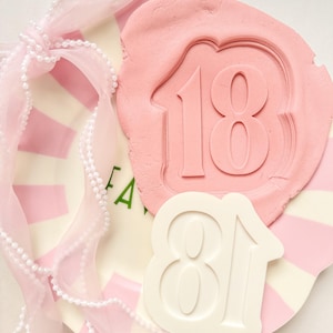 May include: A pink wax seal with the number "18" embossed on it, alongside a white number "81" cutout. A pink and white striped background is visible, with a sheer pink ribbon and pearl trim. The overall theme is celebratory.