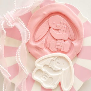 May include: A pink and white striped plate holds a pink bunny-shaped mold and a white bunny-shaped mold. The pink mold has a bunny holding a pencil. A sheer pink ribbon with white beaded trim is tied in a bow.