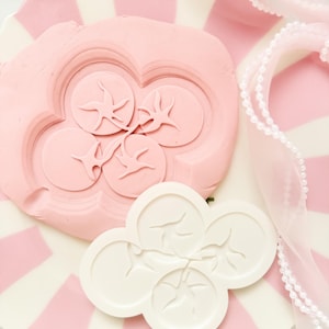 May include: A pink and white striped plate holds a pink floral-shaped mold with a bird design. A white floral-shaped mold with a bird design sits on the plate. A white ribbon with pearls is draped on the plate.