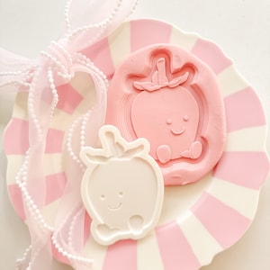 Peut inclure: Une assiette à rayures roses et blanches contient un moule rose en forme de pomme et un objet blanc en forme de pomme, tous deux avec des visages souriants. Un ruban rose transparent avec des perles est noué en nœud. Les articles sont disposés sur une surface blanche.