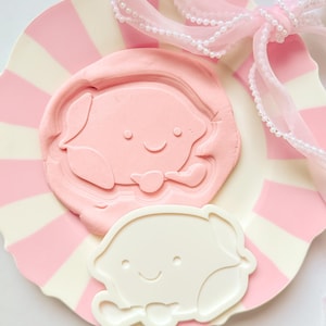Peut inclure: Une assiette à rayures roses et blanches présente un motif de grenouille en relief rose et un emporte-pièce en forme de grenouille blanche. Un ruban rose avec des perles est noué en nœud. La grenouille a un visage souriant.