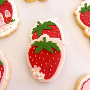 Pode incluir: Um biscoito em forma de morango com glacê vermelho, pequenos detalhes de sementes cor-de-rosa, um caule verde e duas pequenas flores brancas na base, sobre um fundo de glacê rosa claro, rodeado por outros biscoitos decorados.