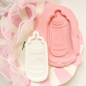 Op de afbeelding: Een witte plastic koekjesvorm in de vorm van een ketchupfles en een roze klei-afdruk van hetzelfde ontwerp, met tomatenillustraties. De items liggen op een roze en wit gestreepte plaat, versierd met een roze lint.