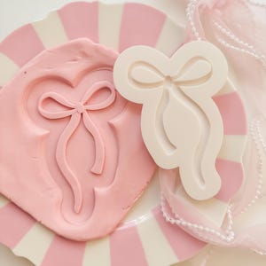 May include: A pink bow-shaped mould and a pink clay impression on a striped plate. A white bow-shaped mould sits next to the clay. A sheer pink ribbon with pearls is draped nearby. The theme is related to crafts and baking.