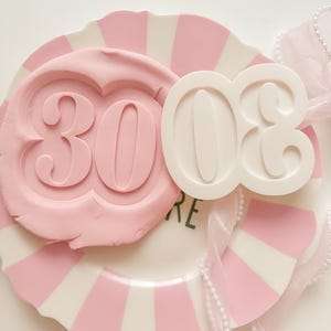 May include: A plate with pink and white stripes, with a pink embossed "30" and a white "03" design. A pink ribbon with pearls adds a delicate touch. The word "RE" is visible in green.